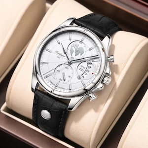 May include: A silver-toned wristwatch with a white face and black leather band. The watch face features multiple dials, including a world map design. The band is textured, and the watch is displayed on a cream-colored surface.