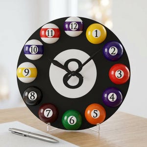 May include: A black round clock with a white number eight in the center. The clock face features billiard balls in place of numbers. The clock hands are black. The clock is displayed on a light-colored surface.
