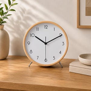 May include: A round analog table clock with a light wood frame and a white face. The clock features black numerals from 1 to 12, black hour and minute hands, and a thin black second hand. It stands on two small, silver-toned metal legs.