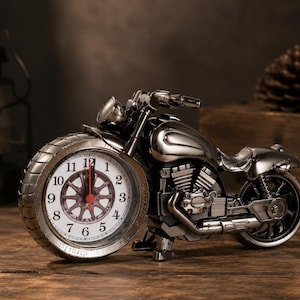 May include: A metallic silver motorcycle-shaped table clock featuring a round white clock face integrated into the front wheel. The clock has black numerals, a red second hand, and detailed engine components, handlebars, and a rear wheel structure.