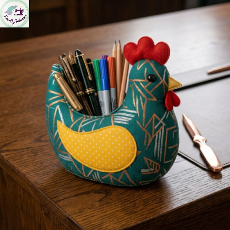 K&ouml;nnte beinhalten: Ein skurriler Stifthalter in Form eines Huhns aus teal-farbenem Stoff mit goldenem geometrischen Muster. Das Huhn hat einen roten Kamm und einen gelben Schnabel. Der Halter ist mit Stiften und Bleistiften gef&uuml;llt. Der Fl&uuml;gel ist gelb mit wei&szlig;en Punkten.