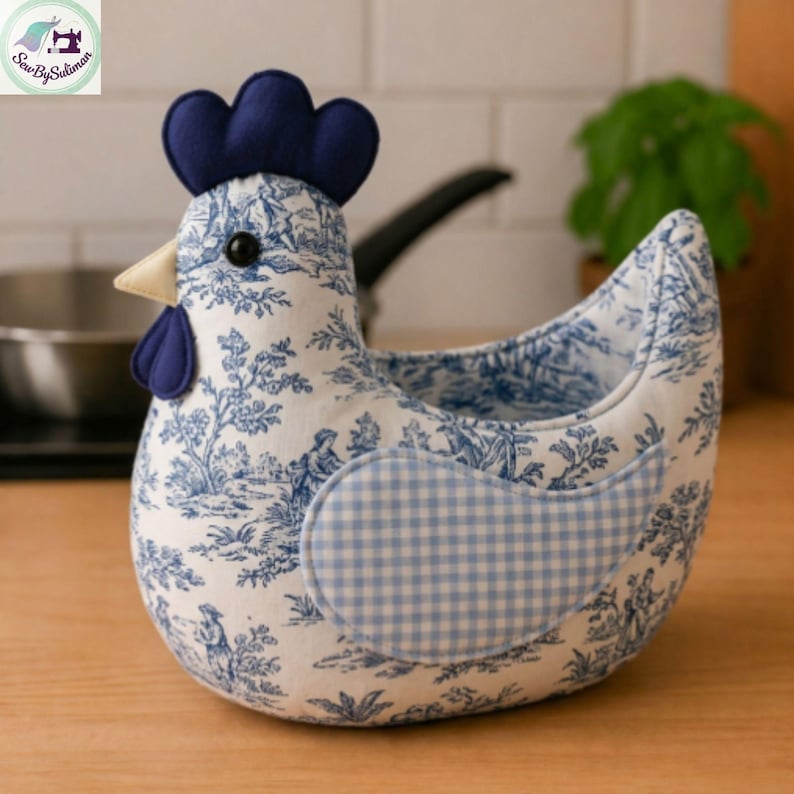 K&ouml;nnte beinhalten: Ein stoffbezogener H&uuml;hnerkorb mit blau-wei&szlig;em Toile-de-Jouy-Muster. Das Huhn hat einen blauen Kamm und Kehllappen, einen gelben Schnabel und einen blau-wei&szlig; karierten Fl&uuml;gel. Der Korb ist zum Aufbewahren von Gegenst&auml;nden gedacht.