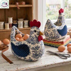 K&ouml;nnte beinhalten: Zwei stoffbezogene Eierhalter in Form eines Huhns. Die Halter sind wei&szlig; mit einem blauen Toile-de-Jouy-Muster und haben rote Kamm- und Kehllappen-Details. Einer ist mit braunen Eiern gef&uuml;llt. Eine Schere und ein Ma&szlig;band liegen auf dem Tisch.