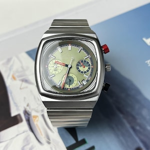 Puede incluir: Un reloj cronógrafo de acero inoxidable con caja cuadrada y esfera verde claro texturizada. El reloj cuenta con tres subesferas, una aguja de cronógrafo roja, un pulsador rojo en la parte superior derecha y un brazalete de eslabones de metal.