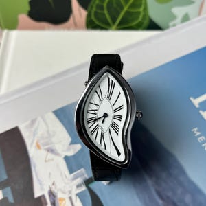 May include: A unique wristwatch with a white face featuring black Roman numerals and black hands. The watch has a silver-colored case and a black leather band. The watch's face has a distinctive, melting shape. The watch is displayed on a blue and white surface.