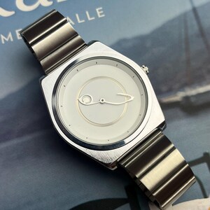 Puede incluir: Reloj de pulsera plateado con esfera cuadrada y blanca. El reloj presenta un diseño minimalista con un logotipo estilizado y una correa de metal. El reloj se muestra sobre un fondo azul y blanco.