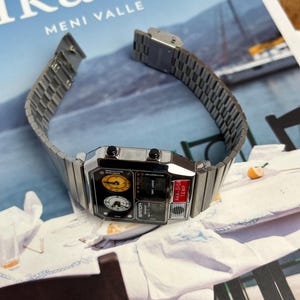 May include: A silver-toned digital watch with a rectangular face and a metal band. The watch face features multiple dials and a digital display. The watch is displayed on a white surface with a blurred background.