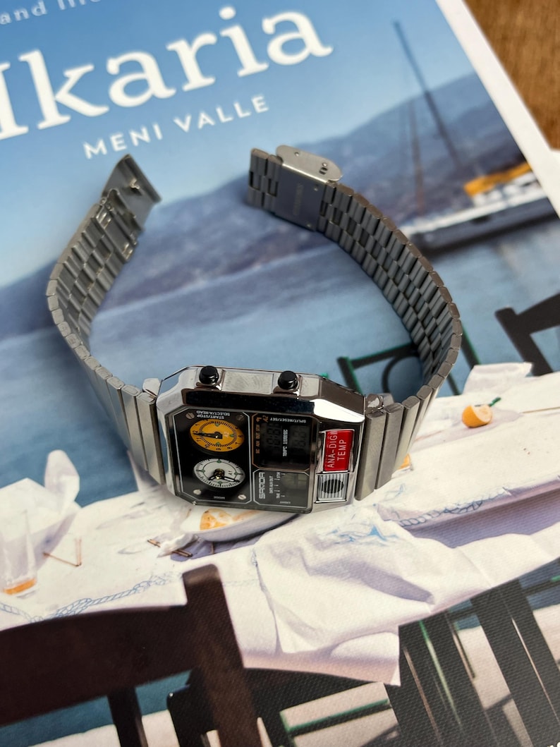 May include: A silver-toned digital watch with a rectangular face and a metal band. The watch face features multiple dials and a digital display. The watch is displayed on a white surface with a blurred background.