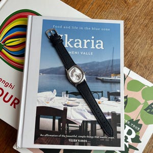 May include: A silver-toned watch with a black leather band rests on a cookbook titled "karia". The book cover features a coastal scene with the text "Food and life in the blue zone". Other books are visible in the background.