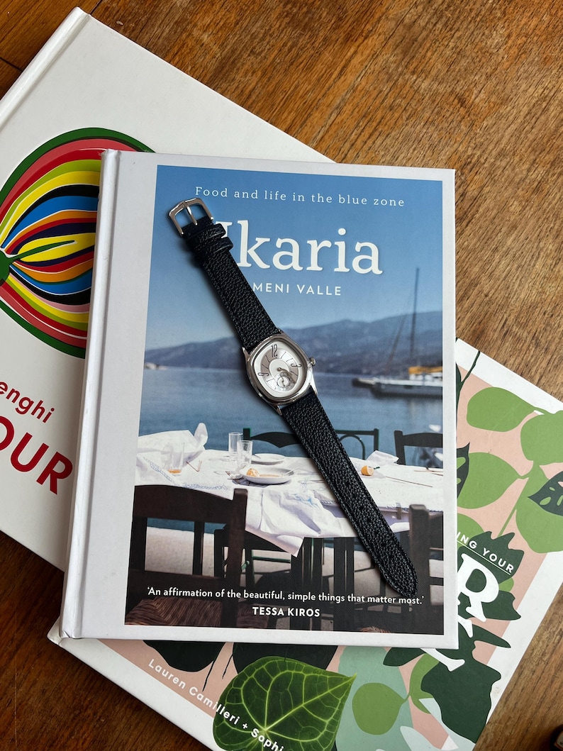 May include: A silver-toned watch with a black leather band rests on a cookbook titled "karia". The book cover features a coastal scene with the text "Food and life in the blue zone". Other books are visible in the background.