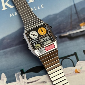 May include: A silver and black digital watch with a rectangular face and a metal band. The watch face displays the time, date, and temperature. The watch has two analog dials and the text "SAMOA" is visible.
