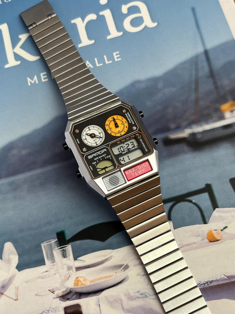 May include: A silver and black digital watch with a rectangular face and a metal band. The watch face displays the time, date, and temperature. The watch has two analog dials and the text "SAMOA" is visible.