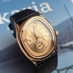 May include: A gold-toned wristwatch with a black leather band. The watch face has a unique oval shape with gold hands and a small seconds dial. The watch has a gold crown and is set against a blue background.