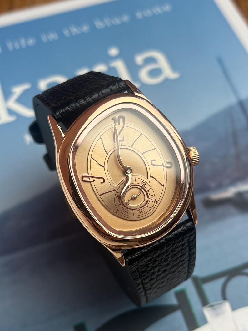 May include: A gold-toned wristwatch with a black leather band. The watch face has a unique oval shape with gold hands and a small seconds dial. The watch has a gold crown and is set against a blue background.