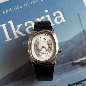 May include: A silver wristwatch with a white face and black leather band. The watch has a unique design with curved numerals and a small seconds dial. The watch is on a blue background with the word "Ikaria" visible.