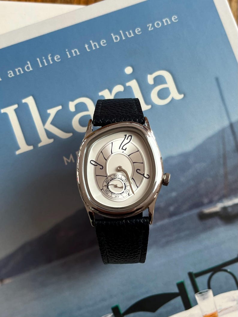 May include: A silver wristwatch with a white face and black leather band. The watch has a unique design with curved numerals and a small seconds dial. The watch is on a blue background with the word "Ikaria" visible.
