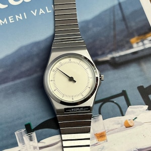 Peut inclure: Une montre-bracelet argent&eacute;e avec un cadran blanc et des aiguilles noires. La montre a un bo&icirc;tier rectangulaire et un bracelet en m&eacute;tal. Le cadran affiche les chiffres de 1 &agrave; 12. La marque "FOSLN" est visible sur le cadran.