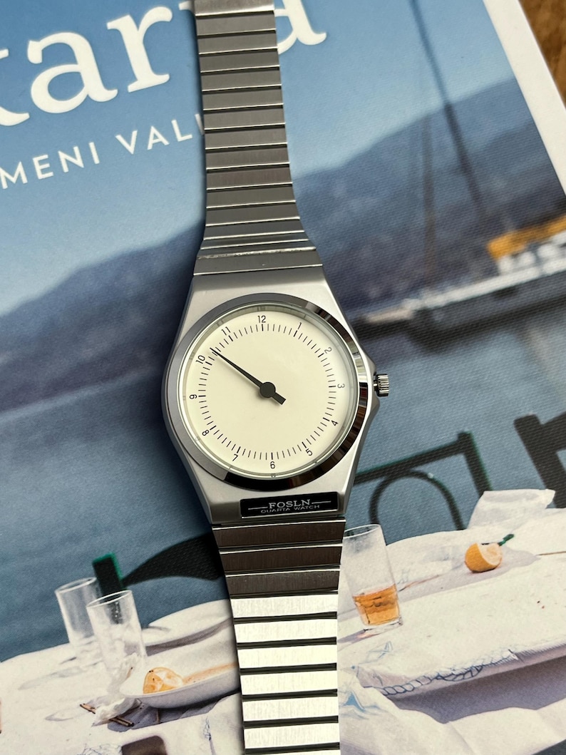 Peut inclure: Une montre-bracelet argent&eacute;e avec un cadran blanc et des aiguilles noires. La montre a un bo&icirc;tier rectangulaire et un bracelet en m&eacute;tal. Le cadran affiche les chiffres de 1 &agrave; 12. La marque "FOSLN" est visible sur le cadran.