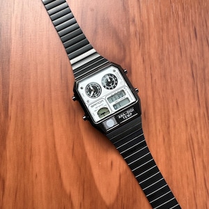 Peut inclure: Une montre-bracelet en m&eacute;tal noir avec un bo&icirc;tier rectangulaire et un bracelet &agrave; maillons assorti. Le cadran pr&eacute;sente deux sous-cadrans analogiques, un affichage num&eacute;rique indiquant 11:39, et le texte SANDA et ANA-DIGI TEMP imprim&eacute;.