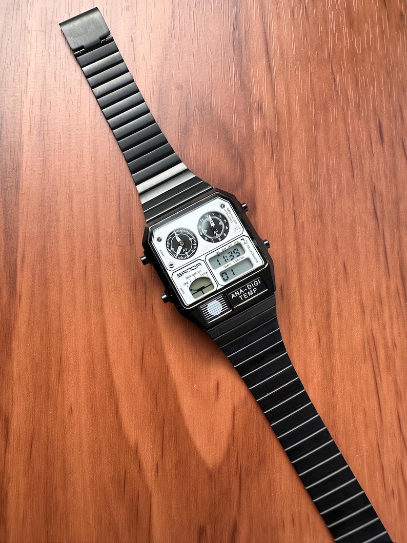 Peut inclure: Une montre-bracelet en m&eacute;tal noir avec un bo&icirc;tier rectangulaire et un bracelet &agrave; maillons assorti. Le cadran pr&eacute;sente deux sous-cadrans analogiques, un affichage num&eacute;rique indiquant 11:39, et le texte SANDA et ANA-DIGI TEMP imprim&eacute;.