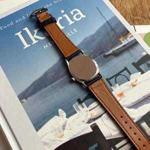 May include: A wristwatch with a brown leather band and a silver-colored watch face. The band has dark blue edges. The watch is on top of a book with the title "Ikaria" and the words "Food and life" visible.