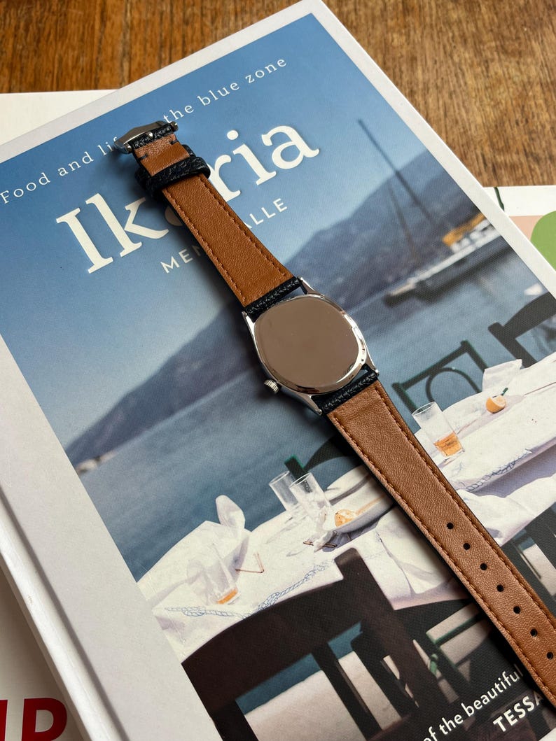 May include: A wristwatch with a brown leather band and a silver-colored watch face. The band has dark blue edges. The watch is on top of a book with the title "Ikaria" and the words "Food and life" visible.