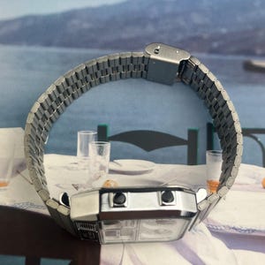 May include: A silver-toned digital watch with a rectangular face and a linked metal band. The watch has two black buttons and a clear display. The band is curved, and the watch is set against a blurred background.