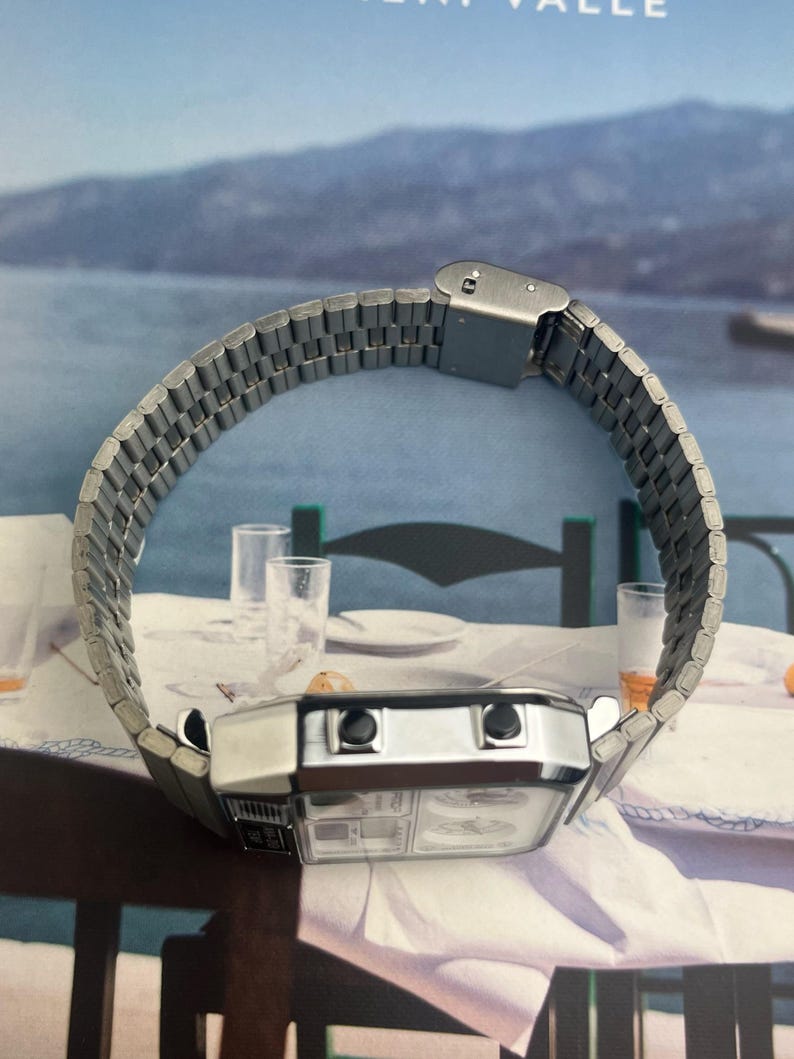 May include: A silver-toned digital watch with a rectangular face and a linked metal band. The watch has two black buttons and a clear display. The band is curved, and the watch is set against a blurred background.
