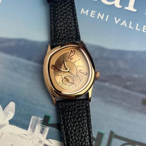 May include: A gold-toned wristwatch with a black leather band. The watch face has a unique shape with a gold dial, featuring the number 12 and a small seconds dial. The band has a textured, pebbled appearance.