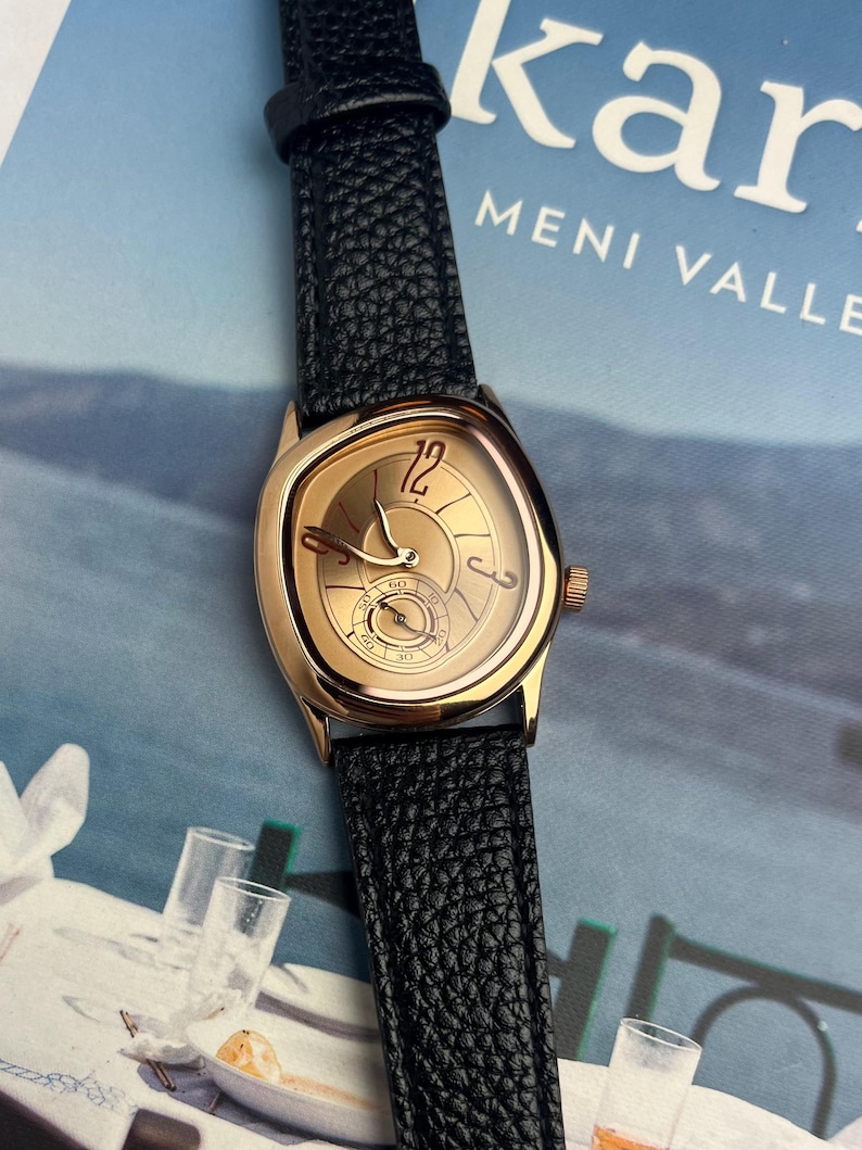 May include: A gold-toned wristwatch with a black leather band. The watch face has a unique shape with a gold dial, featuring the number 12 and a small seconds dial. The band has a textured, pebbled appearance.