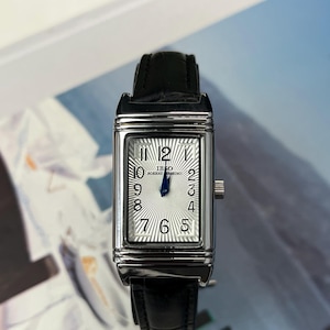 Puede incluir: Un reloj de pulsera rectangular plateado con una correa de cuero negro. La esfera del reloj es blanca con números y agujas negras. El reloj tiene un segundero y la marca "IBSO" es visible.