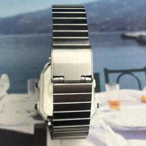 May include: A silver-toned wristwatch with a rectangular face and a metal band. The watch band is composed of multiple rectangular links. The watch is displayed against a blurred background with the text "MENI VALLE".