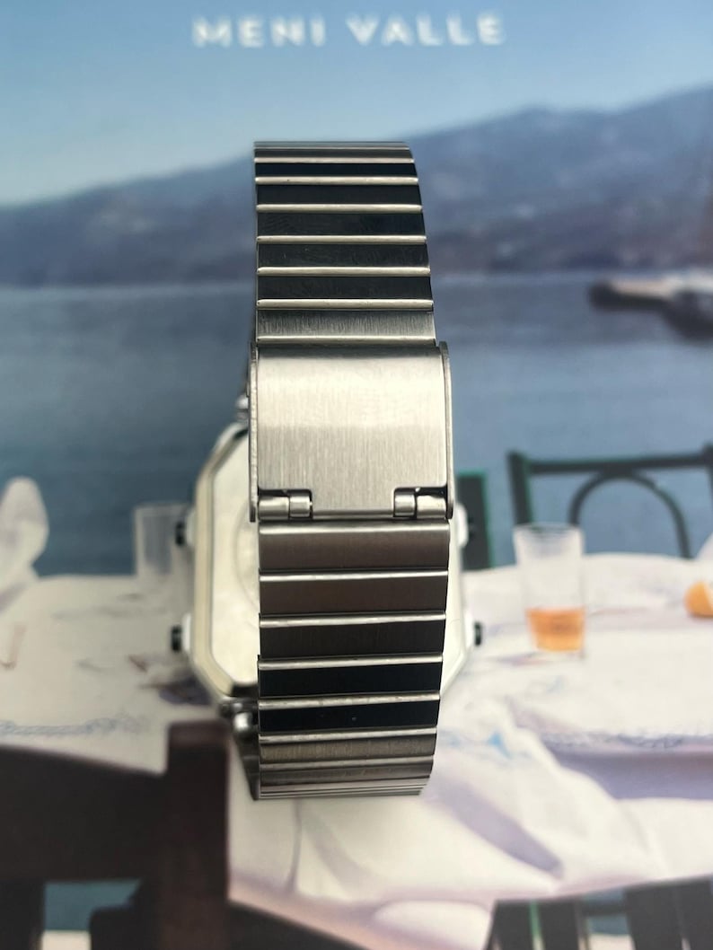 May include: A silver-toned wristwatch with a rectangular face and a metal band. The watch band is composed of multiple rectangular links. The watch is displayed against a blurred background with the text "MENI VALLE".