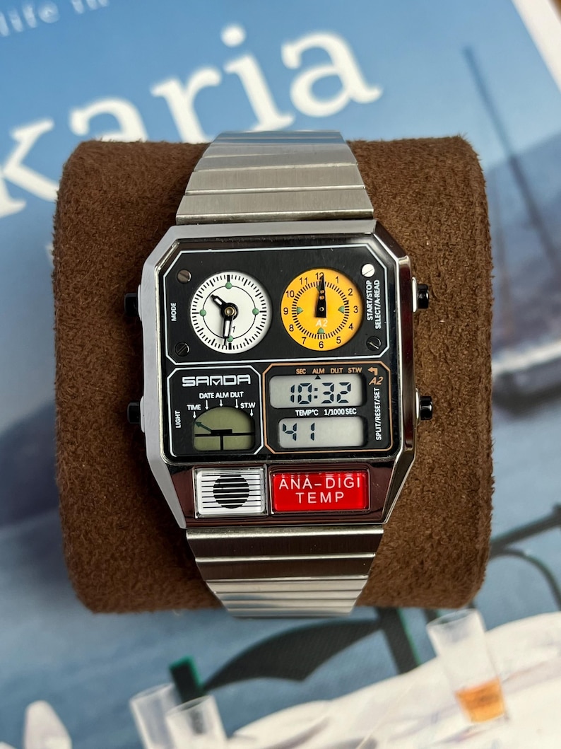 May include: A silver-toned SAMDA digital watch with a black face and a rectangular design. The watch features two analog dials, a digital display showing the time, and the text "ANA-DIGI TEMP" on a red button. The watch is displayed on a brown surface.