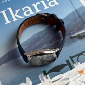May include: A silver wristwatch with a black leather band. The watch face has a detailed design. The watch is resting on a blue surface with the word "Ikaria" printed on it.