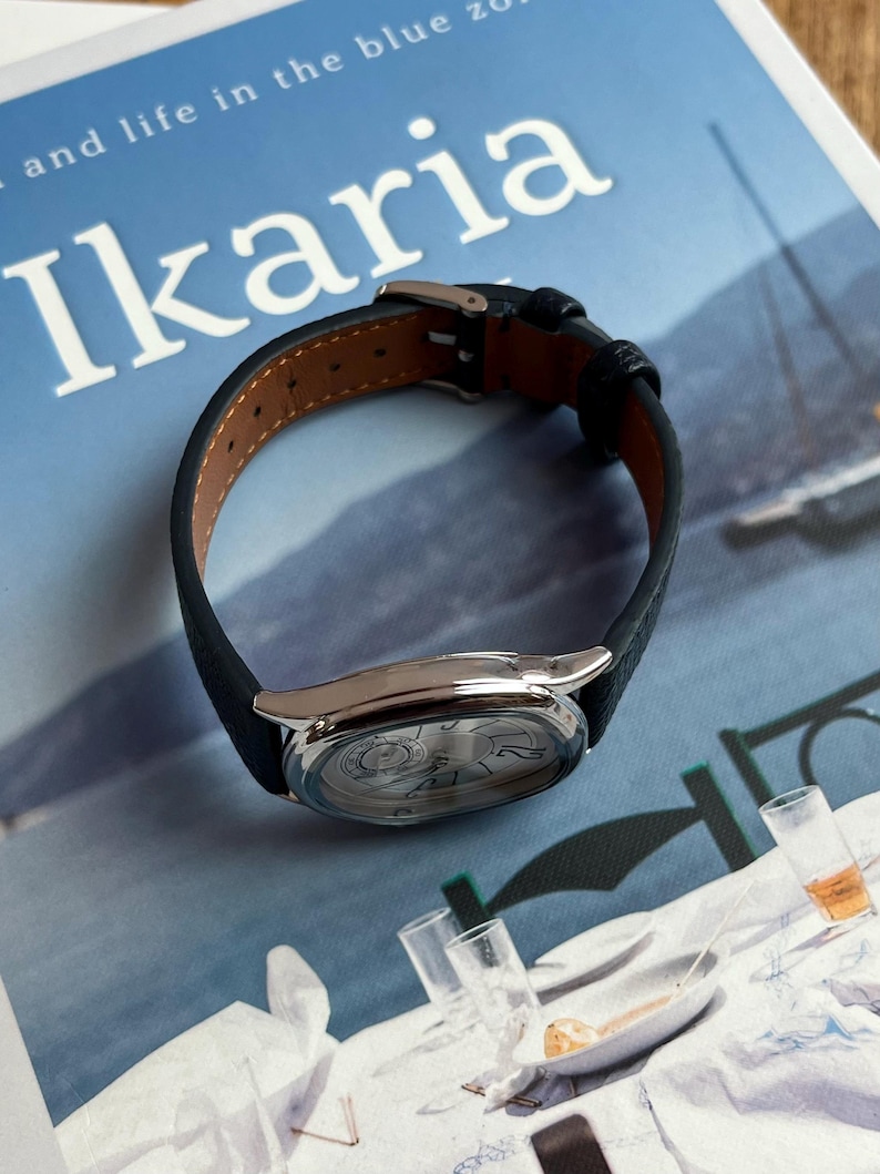 May include: A silver wristwatch with a black leather band. The watch face has a detailed design. The watch is resting on a blue surface with the word "Ikaria" printed on it.