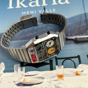 May include: A silver-toned digital watch with a rectangular face and a metal band. The watch face displays multiple dials and digital readouts, including "ANA-DIGI TEMP". The watch is set against a backdrop of a book titled "Ikaria".