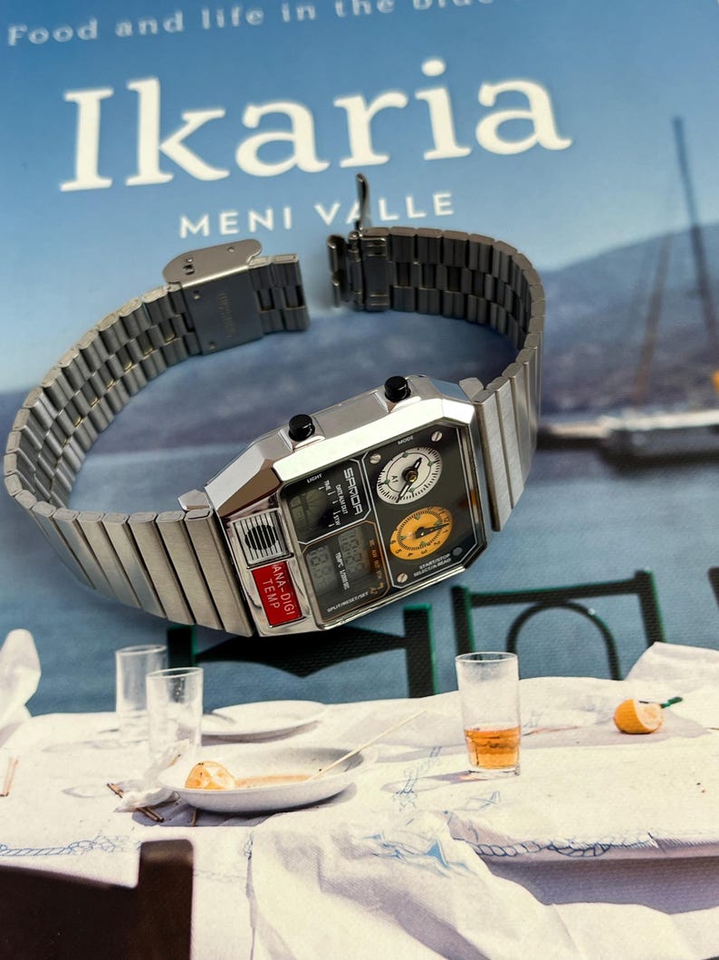 May include: A silver-toned digital watch with a rectangular face and a metal band. The watch face displays multiple dials and digital readouts, including "ANA-DIGI TEMP". The watch is set against a backdrop of a book titled "Ikaria".