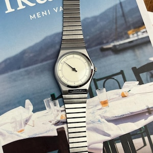 Peut inclure: Une montre-bracelet argent&eacute;e avec un cadran cr&egrave;me et un design minimaliste. La montre pr&eacute;sente un bracelet rectangulaire et un cadran rond avec des aiguilles et des index noirs. Le nom de la marque "FOSLN" est imprim&eacute; sur le bracelet. La montre est pr&eacute;sent&eacute;e sur un magazine.