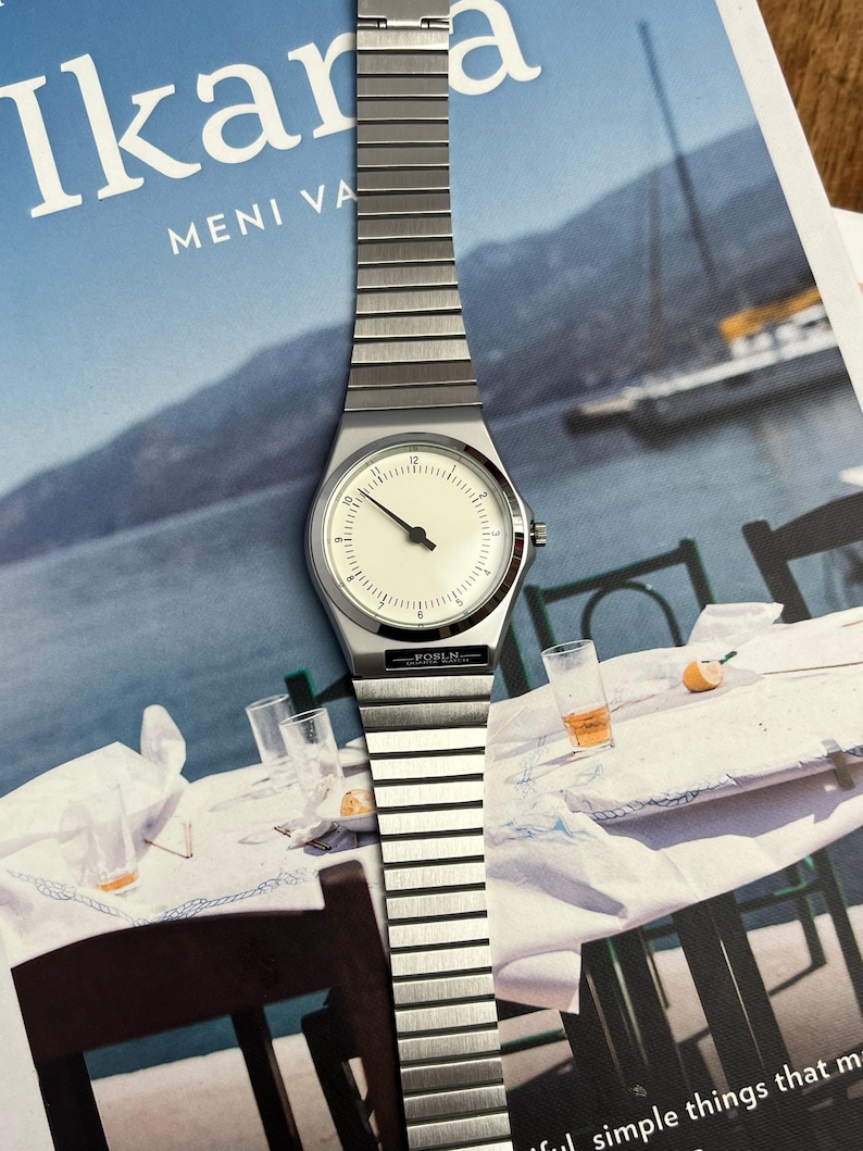 Peut inclure: Une montre-bracelet argent&eacute;e avec un cadran cr&egrave;me et un design minimaliste. La montre pr&eacute;sente un bracelet rectangulaire et un cadran rond avec des aiguilles et des index noirs. Le nom de la marque "FOSLN" est imprim&eacute; sur le bracelet. La montre est pr&eacute;sent&eacute;e sur un magazine.