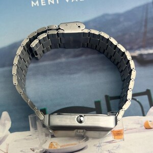 May include: A silver-toned wristwatch with a rectangular face and a linked metal band. The watch is displayed on a table with a book in the background. The text "MENI VALLE" is visible on the book.