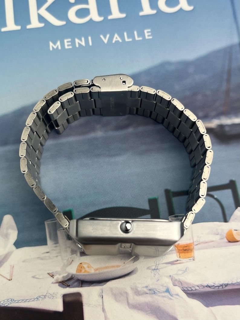 May include: A silver-toned wristwatch with a rectangular face and a linked metal band. The watch is displayed on a table with a book in the background. The text "MENI VALLE" is visible on the book.