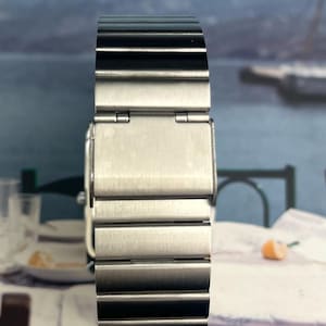 May include: A stainless steel watch band with a brushed metal finish. The band is composed of multiple rectangular links and features a clasp closure. The watch band is designed to be worn on the wrist.