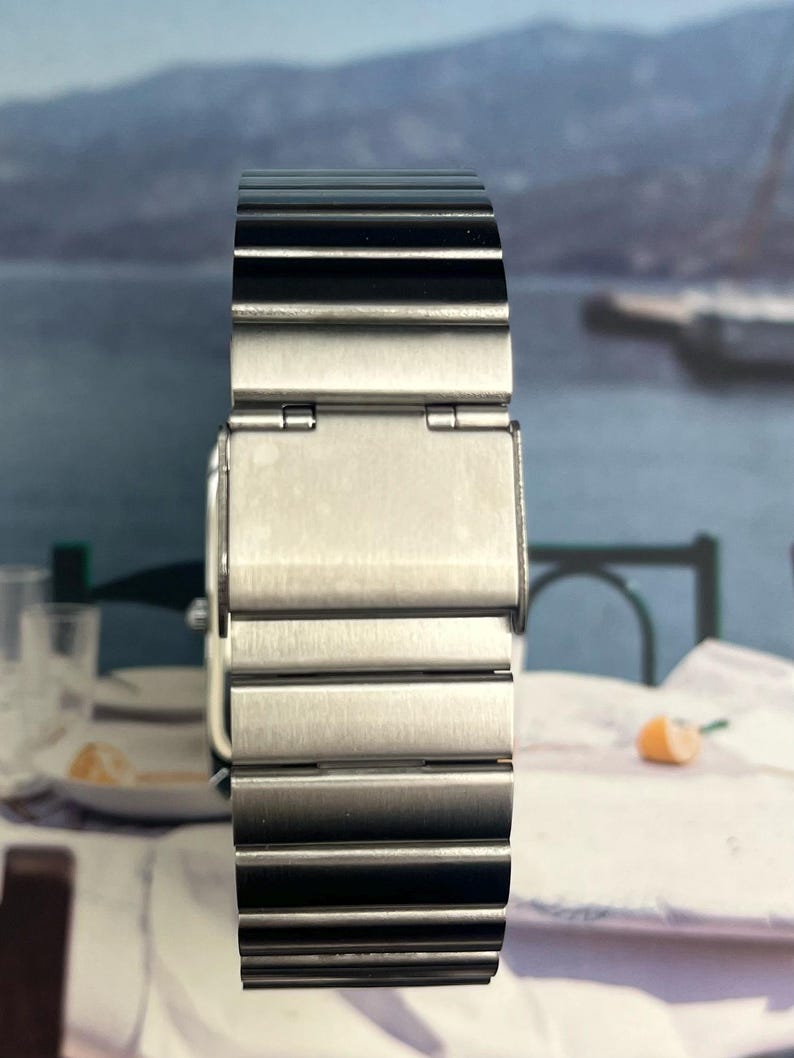 May include: A stainless steel watch band with a brushed metal finish. The band is composed of multiple rectangular links and features a clasp closure. The watch band is designed to be worn on the wrist.