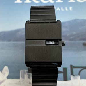 Puede incluir: Un reloj de pulsera negro mate con caja rectangular y correa de eslabones metálicos acanalados a juego. La esfera presenta una abertura horizontal que muestra los dígitos de la hora y una pequeña corona negra en el lado derecho.