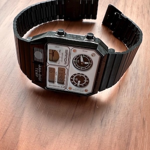 Peut inclure: Une montre-bracelet num&eacute;rique et analogique noire avec un bracelet &agrave; maillons en m&eacute;tal. Le cadran rectangulaire porte l'inscription SAMOA, ANA-DIGI TEMP et diverses &eacute;tiquettes de fonction. Elle comprend deux cadrans analogiques circulaires.