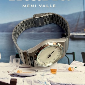 Peut inclure: Une montre-bracelet argent&eacute;e avec un cadran blanc et des aiguilles noires, pos&eacute;e sur une nappe blanche. La montre a un bracelet en m&eacute;tal et le texte "-ROSLN-" est visible. En arri&egrave;re-plan, un livre avec le titre "Ikaria" et le nom "MENI VALLE".