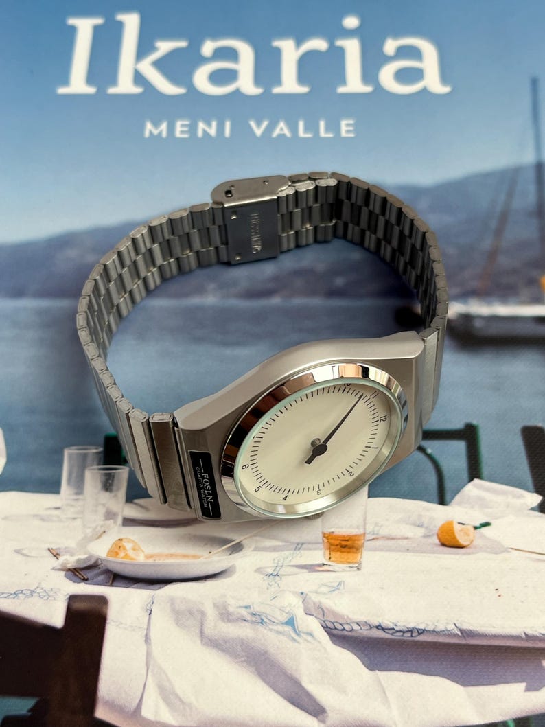 Peut inclure: Une montre-bracelet argent&eacute;e avec un cadran blanc et des aiguilles noires, pos&eacute;e sur une nappe blanche. La montre a un bracelet en m&eacute;tal et le texte "-ROSLN-" est visible. En arri&egrave;re-plan, un livre avec le titre "Ikaria" et le nom "MENI VALLE".