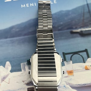 May include: A silver-toned digital watch with a rectangular face and a stainless steel band. The watch is displayed on a table with a blue background and the word "Ikaria" visible.