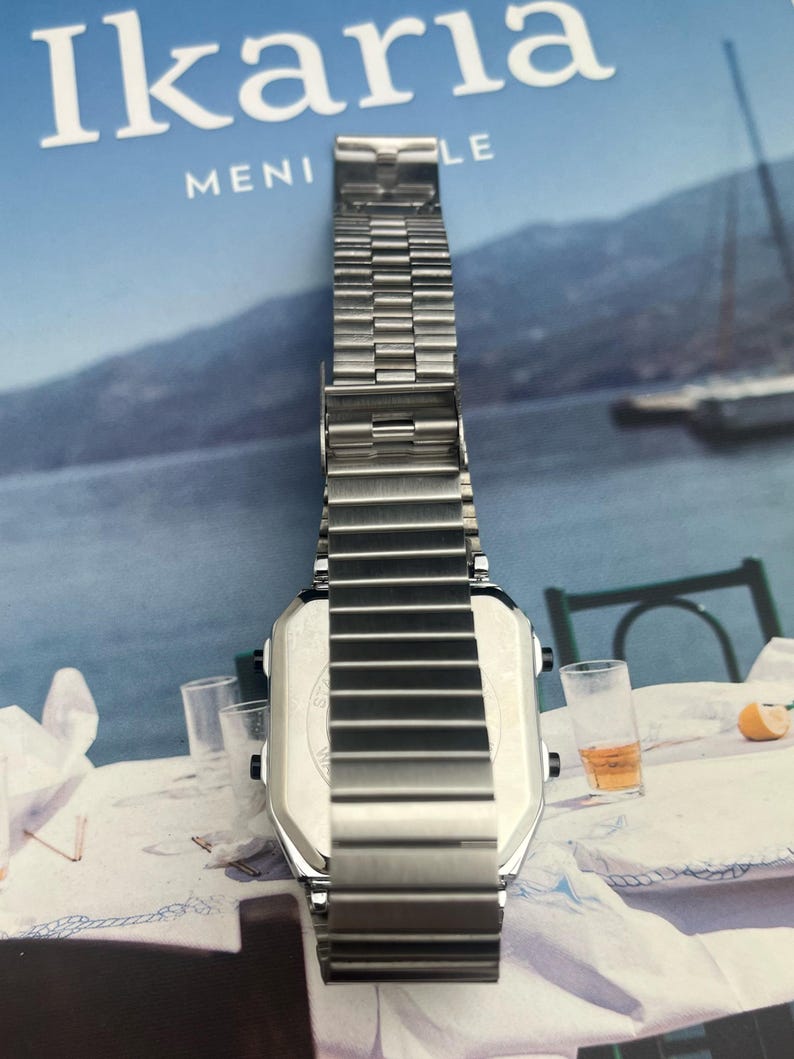 May include: A silver-toned digital watch with a rectangular face and a stainless steel band. The watch is displayed on a table with a blue background and the word "Ikaria" visible.